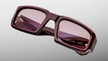 A pair of angular square sunglasses with a glossy dark burgundy frame and tinted pink lenses, shown on a light gray gradient background.