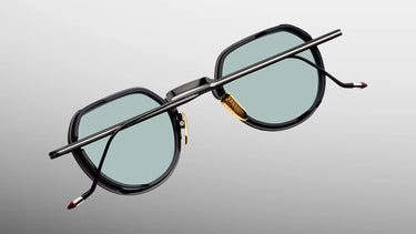 A pair of round eyeglasses with a black frame, light blue-green lenses, and gold metal accents, shown floating against a gray gradient background.