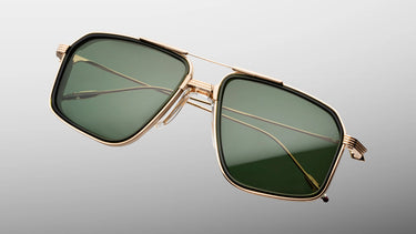A pair of oversized square sunglasses with a gold-tone metal frame and dark green lenses, shown floating against a light gray gradient background.